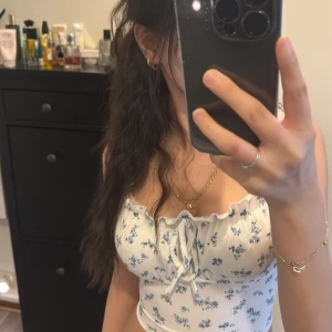 Blommig vit och blå linnetopp - Söt blommig linnetopp i vitt med blå blommor. Toppen har knytning framtill och volangdetaljer vid axelbanden. Perfekt för varma dagar.