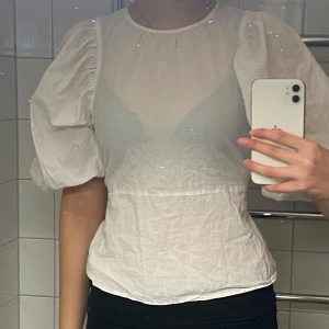 Vit blus med puffärmar - Säljer en stilren vit blus med puffiga kortärmar och en öppen rygg. Blusen har knytband i ryggen för en justerbar passform. Perfekt för en chic och luftig look.