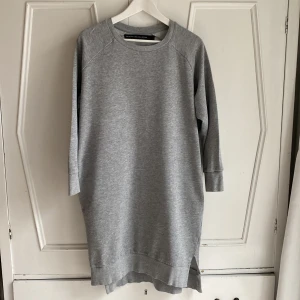 Gråmelerad klänning från The Black Label Collection - Gråmelerad klänning/långtröja från The Black Label Collection. Påsytt svart kryss på ryggen. Små sprund längst ner och fickor i sidorna. Längd 100 cm. 