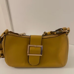 Gul axelväska - Snygg gul axelväska med ett stort metallspänne på framsidan. Väskan har en dragkedja upptill och en justerbar axelrem. Perfekt för att ge en färgklick till din outfit!