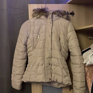 Beige dunjacka från H&M - Säljer en snygg beige dunjacka från H&M med pälskantad huva. Jackan har dragkedja och knappar framtill samt två praktiska fickor med dragkedja. Perfekt för kyliga dagar!