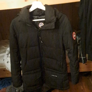 Svart dunjacka från Canada Goose - Säljer en svart dunjacka från Canada Goose med huva och dragkedja. Jackan har ett märke på ärmen och är perfekt för kalla dagar. Den är långärmad och har en stilren design.