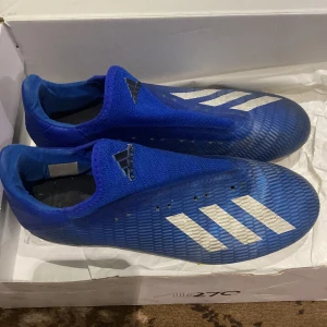 Blå fotbollsskor från Adidas - Snygga blå fotbollsskor från Adidas med vita ränder och en strömlinjeformad design. Skorna har en tight passform och är perfekta för snabba rörelser på planen. De har en låg profil och är utan snörning för en smidig känsla.