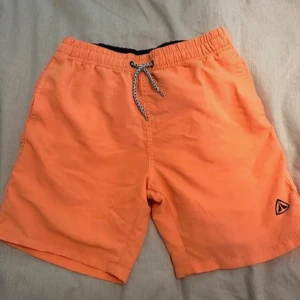 Orange badshorts med snörning - Snygga orange badshorts med elastisk midja och snörning framtill. Perfekta för sommarens alla badäventyr. De har en liten logga på benet som ger en sportig touch.