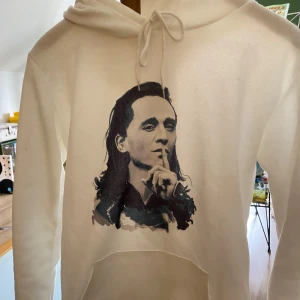 Vit hoodie med tryck - Säljer en vit hoodie med ett tryck av Loki som håller fingret mot läpparna. Hoodien har en klassisk design med dragsko i huvan och en stor ficka framtill. Oklar storlek men passar mig som XS