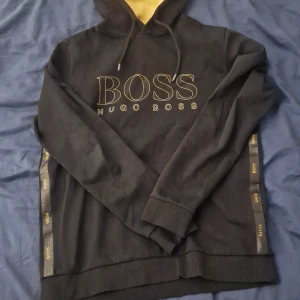 Svart hoodie från Hugo Boss - Snygg svart hoodie från Hugo Boss med gul text och detaljer. Den har en justerbar huva med gul insida och långa ärmar. Perfekt för en stilren och bekväm look. Max 550. Storlek L men passar m