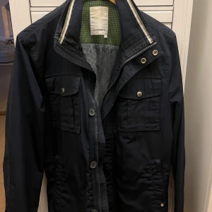 Mörkblå jacka från J. Lindeberg - Snygg svart jacka från J. Lindeberg med dragkedja och knappar framtill. Jackan har flera praktiska fickor med knappar och en stilren design. Perfekt för en stilmedveten look.