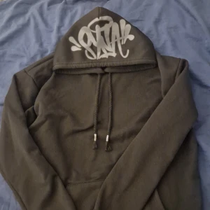 Syna world Svart hoodie med tryck - Säljer en syna world svart hoodie med ett coolt tryck på huvan. Den har en justerbar dragsko och långa ärmar. Perfekt för en avslappnad stil. 300 sista pris
