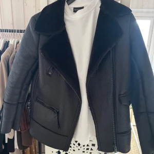 Svart jacka med pälsdetaljer - Snygg svart jacka från Zara med mjuka pälsdetaljer på kragen och insidan. Jackan har en cool bikerstil med dragkedja och fickor. Perfekt för att hålla stilen i kallare väder.