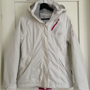 Vit damskidjacka från Helly Hansen, strl M - Säljer en snygg vit skidjacka från Helly Hansen med dragkedja och avtagbarhuva. Jackan har snölås, en praktisk ficka med dragkedja på bröstet och är utrustad med RECCO-reflektor för ökad säkerhet. Jackan är vind-och vattentät.
