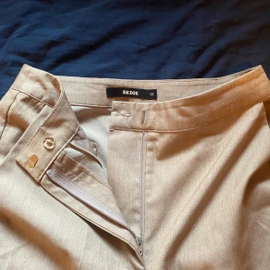 Beige kostymbyxor från Bik Bok - Snygga beige kostymbyxor från Bik Bok i storlek 36. De har en stilren design med dragkedja och knapp framtill. Byxorna har en rak passform, lite utsvängda där mete och är perfekta för en chic look. Materialet känns mjukt och bekvämt.
