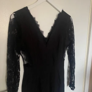 Svart jumpsuit med spetsdetaljer - Elegant svart jumpsuit med långa spetsärmar och v-ringning. Perfekt för en stilren look med fina spetsdetaljer som ger en extra touch. Passar bra till både formella och avslappnade tillfällen.