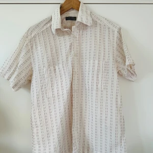 Vit och brun randig skjorta från The Shirt Factory - Snygg kortärmad skjorta från The Shirt Factory designad av Linnea Braun. Skjortan är vit med tunna röda ränder och har knappar framtill. Perfekt för en stilren och avslappnad look.