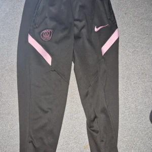 Svarta träningsbyxor med rosa detaljer - Snygga svarta träningsbyxor från Nike med rosa detaljer och PSG-logga. Perfekta för sport och träning med en bekväm passform och elastisk midja.