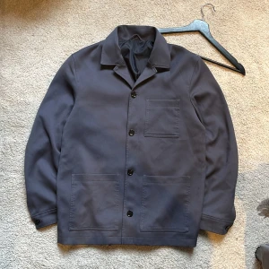 Overshirt Grå Storlek M/L  - Snygg mörkgrå overshirt från H&M med klassisk krage och fyra knappar framtill. Jackan har två stora fickor och en bröstficka. Perfekt för en stilren look.