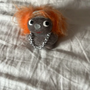 Grått stenhalsband med orange hår - Unikt handgjort halsband med en grå stenfigur som har busigt orange hår. Figuren har stora ögon och ett litet leende, vilket ger den en charmig karaktär. Kedjan är i silverfärg och ger en cool kontrast till den lekfulla designen.