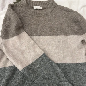 Grå och beige randig tröja - Säljer en mysig tröja i grå och beige med breda ränder. Tröjan har en rund hals och långa ärmar. Tyvärr lite nopprig men annars super fin