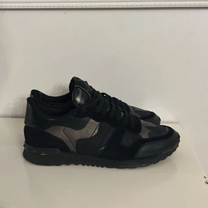 Svarta sneakers från Valentino - Snygga svarta sneakers från Valentino med ett diskret camouflage-mönster. Skorna har snörning och är tillverkade i en kombination av skinn och syntetmaterial. Perfekta för en stilren look äkta bra skick stolek 40
