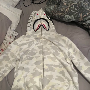 Vit camo hoodie från A Bathing Ape - Säljer en cool vit camo hoodie från A Bathing Ape med det ikoniska hajmotivet på huvan. Hoodien har dragkedja och ribbade muddar. Perfekt för streetwear-entusiaster! Om du har frågor så skriv gärna