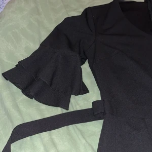 Svart jumpsuit med volangärmar - Elegant svart jumpsuit med v-ringning och volangärmar. jumpsuiten har ett knytband i midjan för en smickrande passform. Perfekt för en stilren look.