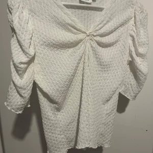 Vit blus med struktur - Elegant vit blus med v-ringning och trekvartsärm. Blusen har en unik textur och ett knytliknande detalj framtill som ger en stilfull look. Perfekt för att lyfta vilken outfit som helst.