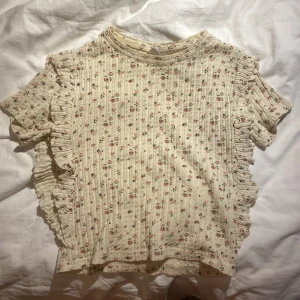 Blommig ribbad topp - blommig topp med ribbad struktur och volangdetaljer. Den har korta ärmar och en mjuk, krämfärgad bas med små rosa blommor. 