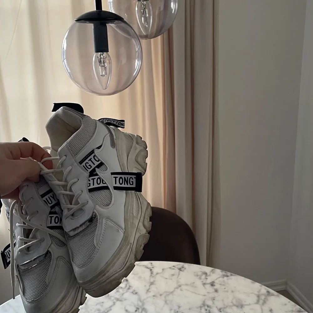 Snygga vita sneakers med tjock sula och textdetaljer i svart och vitt. Skorna har snörning och en modern design som passar perfekt för en trendig look.. Kengät.