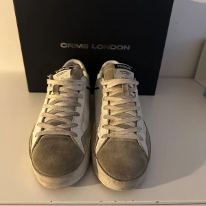 Vita sneakers från Crime London - Snygga vita sneakers från Crime London med grå detaljer och snörning. Skorna har en stilren design med mörkblå häl och märkets logga på tungan och hälen. Perfekta för en avslappnad stil. Skorna är i storlek 39. Box fås med 