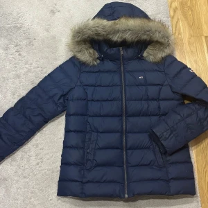 Blå dunjacka från Tommy Jeans - Snygg blå dunjacka från Tommy Jeans med pälskantad huva och dragkedja. Perfekt för kalla dagar med sin quiltade design och långa ärmar. Jackan har märkets logga på bröstet och ärmen.