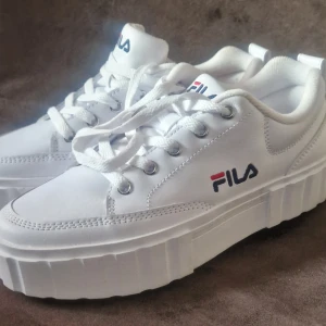 Vita sneakers från Fila - Oanvända snygga vita sneakers från Fila med en chunky sula och klassisk snörning. Perfekta för en stilren look med Fila-loggan i rött och blått på sidan och tungan.