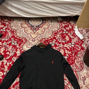 Svart hoodie från Ralph Lauren - Säljer en stilren svart hoodie från Ralph Lauren med en liten röd logga på bröstet. Tröjan har långa ärmar och en klassisk huva med dragsko. Perfekt för en avslappnad look.