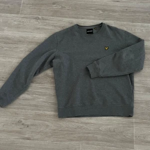 Grå sweatshirt från Lyle & Scott - Snygg grå sweatshirt från Lyle & Scott med deras ikoniska logga på bröstet. Tröjan har långa ärmar och en klassisk rund halsringning. 
