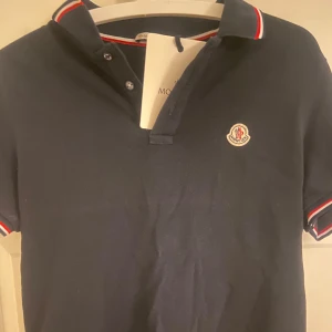 Mörkblå pikétröja från Moncler - Säljer en stilren mörkblå pikétröja från Moncler med klassisk krage och knappar. Tröjan har korta ärmar och Monclers logotyp broderad på bröstet. Skriv vid minsta intresse