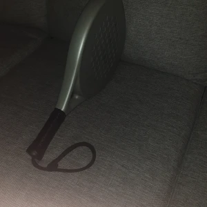 Grå padelracket - Oanvänd padelrack köpt 2024 sommaren.  Säljer en stilren grå padelracket med rund form och perforerad yta. Racketen har ett svart grepp och handledsrem för extra säkerhet. Perfekt för både nybörjare och erfarna spelare som vill ha en pålitlig racket på banan. NY PRIS ÄR 500 MITT PRIS 350 pris kan diskuteras pm.