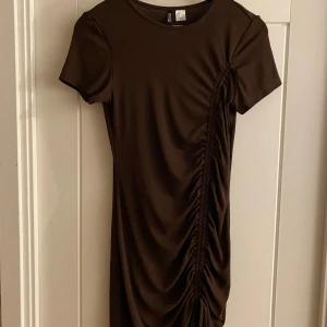 Brun klänning från H&M Divided - Snygg brun klänning från H&M Divided med korta ärmar och rynkdetaljer längs sidan. Perfekt för en stilren look.