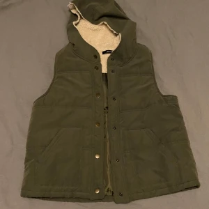 Grön väst med huva från Begun - Grön väst med huva. Lurvigt tyg på insidan, matt på utsidan. Stängs med tryckknappar. Rak passform 