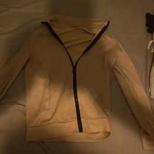 Beige huvtröja med dragkedja - Säljer en beige huvtröja med dragkedja och långa ärmar. Perfekt för en avslappnad stil. Tröjan har en enkel design med en bekväm passform.