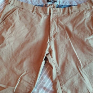 Beige chinos från Dressmann - Snygga beige chinos från Dressmann med regular fit. Byxorna har en klassisk design med knappar och bälteshällor. Perfekta för en stilren look.