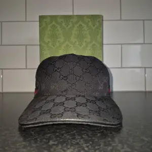 Snygg svart keps från Gucci med det ikoniska monogrammönstret. Kepsen har en röd och grön randdetalj på sidan. Perfekt för att ge din outfit en lyxig touch. Nyskick endast testad. Box och dustbag medföljer
