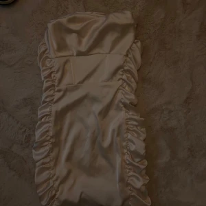 Champagnefärgad klänning från Oh Polly - Elegant champagnefärgad klänning från Oh Polly i satin. Klänningen har en åtsittande passform med volangdetaljer längs sidorna och en dragkedja bak. Perfekt för en glamorös kväll.