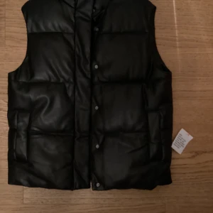 Svart dunväst från ASOS DESIGN - Snygg svart dunväst från ASOS DESIGN med knappar framtill. Perfekt för lager-på-lager under kyliga dagar. Västen har en stilren design och är lätt att matcha med olika outfits.