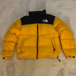 Gul och svart dunjacka från The North Face - Säljer en snygg gul och svart dunjacka från The North Face. Jackan har en dragkedja framtill och är långärmad med justerbara ärmslut. Perfekt för kalla dagar med sin pufferdesign.