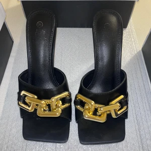 Svarta sandaler med gulddetaljer - Eleganta svarta sandaler med en stilren design och guldfärgade kedjedetaljer framtill. Sandalerna har en smal klack och en fyrkantig tå som ger en modern touch. Perfekta för att lyfta vilken outfit som helst. Obs: Använda en gång! Storlek 36, köpta i Stockholm (Yes London)!✨