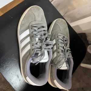 Grå Adidas sneakers med vita ränder - Snygga grå sneakers från Adidas med klassiska vita ränder på sidan. Skorna har en stilren design med snörning och en bekväm sula. Perfekta för en avslappnad stil. Skostorlek 46.