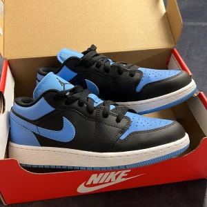 Nike Jordan 1 Low i svart och blått - Snygga Nike Jordan 1 Low i en fräsch kombination av svart och blått (university blue). Skorna har en klassisk design med snörning och en bekväm passform. Nyskick då de enbart är använda en gång. Storlek 37,5 och är svåra att få tag på, inköpta från Nikes egna hemsida. Tillhörande kartong medföljer.