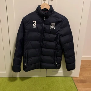 Svart pufferjacka från Vinson Polo Club - Säljer en svart pufferjacka från Vinson Polo Club med broderad logga och nummerdetaljer på framsidan. Jackan har en hög krage och dragkedja framtill. Perfekt för kyliga dagar.