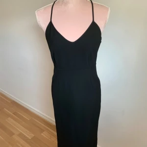 Svart klänning med öppen rygg - Elegant svart klänning med smala axelband och en stilren v-ringning. Klänningen har en vacker öppen rygg med transparenta detaljer som ger en sofistikerad look. Perfekt för en kväll ute eller en speciell tillställning.