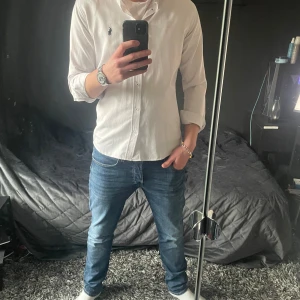 Vit skjorta från Polo Ralph Lauren - Snygg vit skjorta från Polo Ralph Lauren med klassisk krage och broderad logga i blått på bröstet. Perfekt för en stilren look. Skjortan har långa ärmar och knappar framtill. Grabben på bilden är 189 80kg