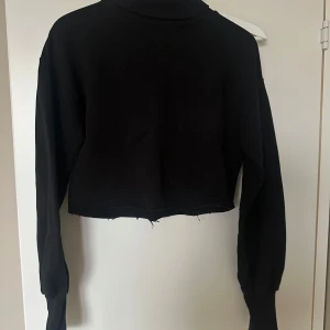 Svart croppad polotröja, S - Säljer en svart croppad tröja med långa ärmar. Tröjan har en enkel design med ribbade muddar vid ärmsluten. Perfekt för en stilren look. Ser avklippt ut (men den är köpt så på Nelly) så lite slitna kanter längst ner för en cool look. 