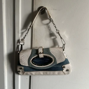  axelväska i jeans och reptilmönster - Snygg axelväska i blått jeansmaterial med vita reptilmönstrade detaljer. Väskan har en unik oval öppning och en kedja som axelrem. Perfekt för att ge din outfit en cool touch. Inköpt 2024 i Italien. Använd en gång.